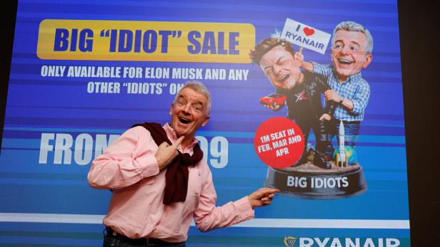 Ein Mann posiert lachend vor einer Werbetafel von Ryanair mit einer Karikatur und dem Schriftzug „BIG IDIOT SALE“.