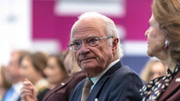 König Carl XVI. Gustaf mit Brille und Anzug sitzt neben Königin Silvia bei einer Veranstaltung.