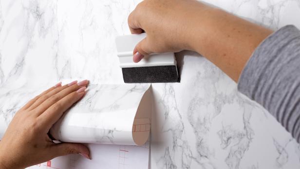 Hände bringen eine Tapete in Marmoroptik mit einer Rakel an einer Wand an.