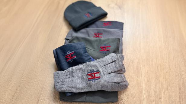 Mütze, Handschuhe und T-Shirts mit gesticktem roten und blauen Logo auf Holztisch.