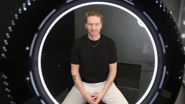 Ein Mann mit Tattoos sitzt vor einer leuchtenden Ringlampe und schaut direkt nach vorne.