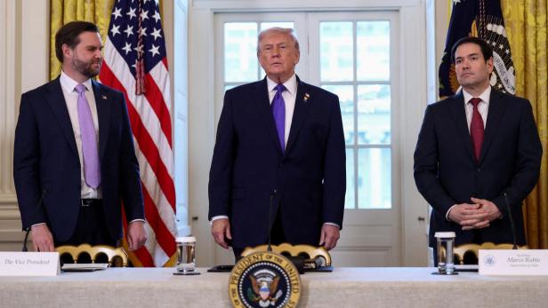 Donald Trump steht mit zwei Männern, JD Vance und Marco Rubio, vor US-Flaggen.