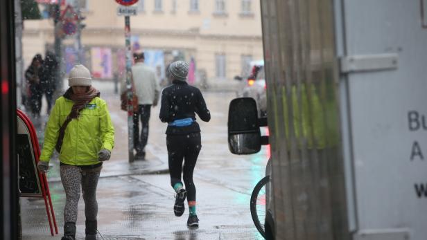 Mehrere Menschen gehen und joggen bei nassem, kaltem Wetter durch eine Stadtstraße, während es leicht schneit.
