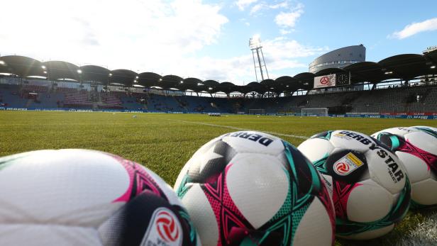 Mehrere Fußbälle liegen am Spielfeldrand eines leeren Fußballstadions bei Tageslicht.