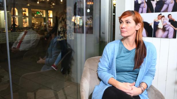 Eine Frau mit rotbraunen Haaren sitzt auf einem Sessel, im Hintergrund sind Fotos und eine Glaswand mit Spiegelung.