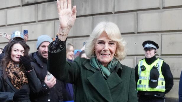 Camilla, in einem grünen Mantel, winkt einer Menschenmenge vor einem Gebäude zu.