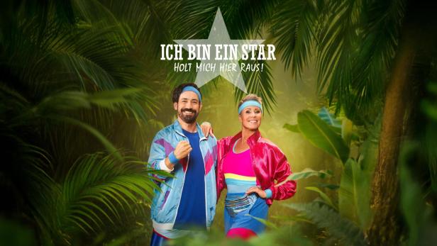 Jan Köppen und Sonja Zietlow in bunter Sportkleidung posieren im Dschungel vor dem Show-Logo.