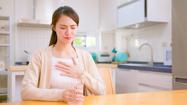 Eine Frau sitzt in einer Küche, hält ein Glas Wasser und fasst sich mit schmerzverzerrtem Gesicht an die Brust.