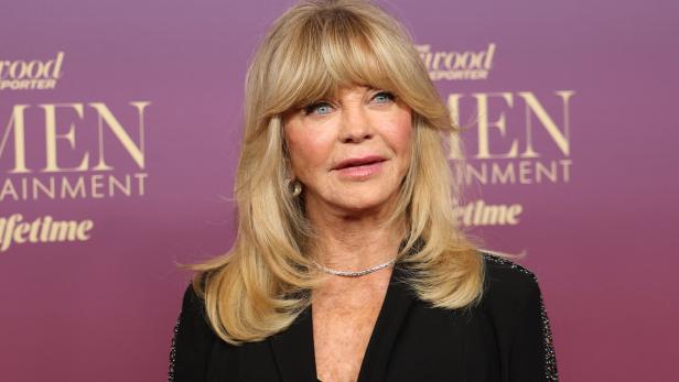 Goldie Hawn mit blondem, schulterlangem Haar und Pony steht vor einem lila Hintergrund mit Schriftzug.