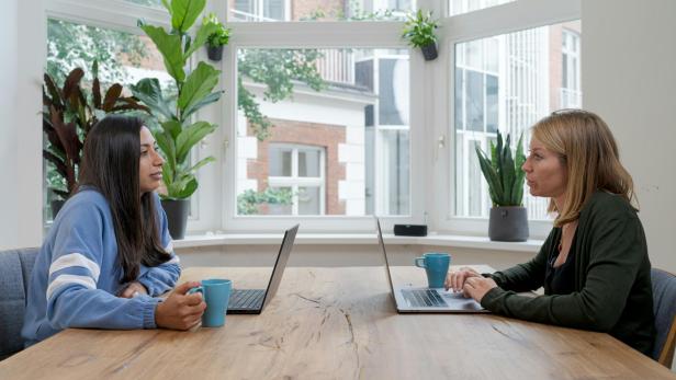 Zwei Frauen sitzen sich an einem Tisch mit Laptops gegenüber und besprechen Content-Marketing-Strategien in einem modernen Büro mit großen Fenstern und Pflanzen.