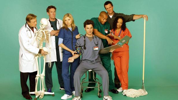 Sieben Personen in Krankenhauskleidung stehen zusammen, eine Person sitzt, ein Skelett und ein Wischmopp sind zu sehen.