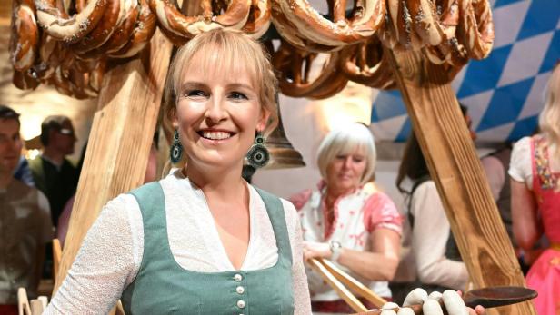 Eine Frau in Tracht hält eine große Holzstange mit Weißwürsten, im Hintergrund hängen viele Brezeln.