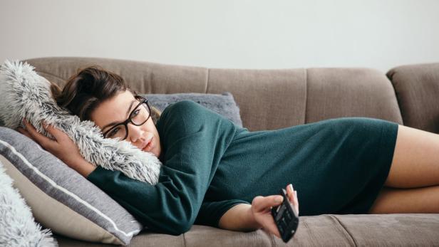 Eine Frau mit Brille liegt entspannt auf einem Sofa, hält eine Fernbedienung und kuschelt sich an ein flauschiges Kissen.