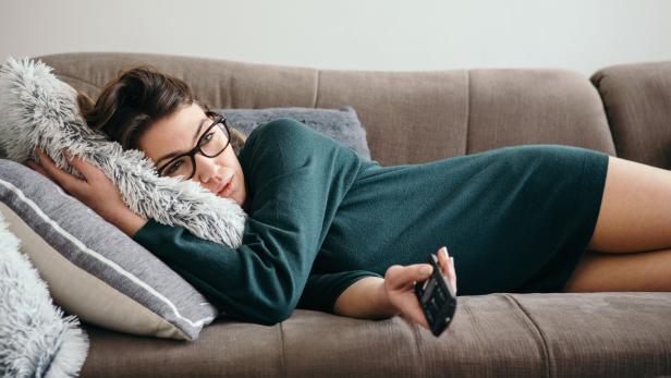 Eine Frau mit Brille liegt entspannt auf einem Sofa, hält eine Fernbedienung und kuschelt sich an ein flauschiges Kissen.