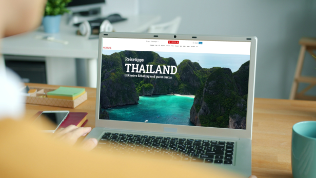 Eine Person betrachtet am Laptop eine digitale Magazinseite des KURIER mit Reisetipps für Thailand und tropischer Landschaft im Hintergrund.