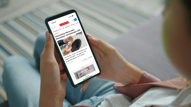 Person hält Smartphone mit geöffneter KURIER-Website und liest einen Artikel über Glasrecycling.