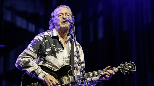 Ein älterer Mann - Peter Cornelius - mit Brille spielt E-Gitarre und singt auf einer Bühne vor dunklem Hintergrund.