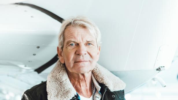 Ein älterer Mann mit grauen Haaren und Fliegerjacke steht vor einem weißen Flugzeug.