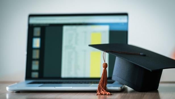 Ein Laptop steht auf einem Tisch, daneben liegt ein schwarzer Doktorhut mit Quaste.
