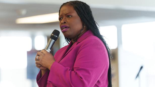 Frau mit langen Zöpfen und pinkem Blazer spricht mit Mikrofon in der Hand vor unscharfem Hintergrund.