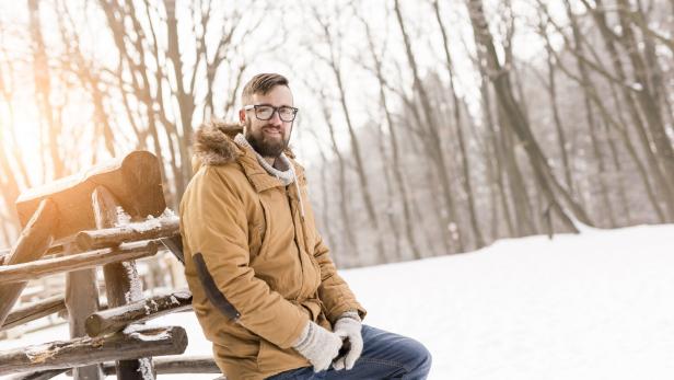 Ein Mann mit Brille und Winterjacke sitzt auf einem Holzzaun im verschneiten Wald.