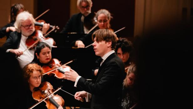 Ein Dirigent - Klaus Mäkelä -  leitet ein Orchester, dessen Musiker konzentriert Streichinstrumente spielen.