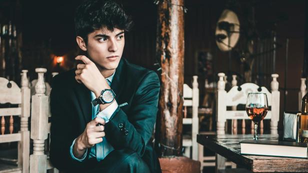 Junger Mann im Anzug sitzt nachdenklich in einem rustikalen Restaurant neben einem Glas Wein.