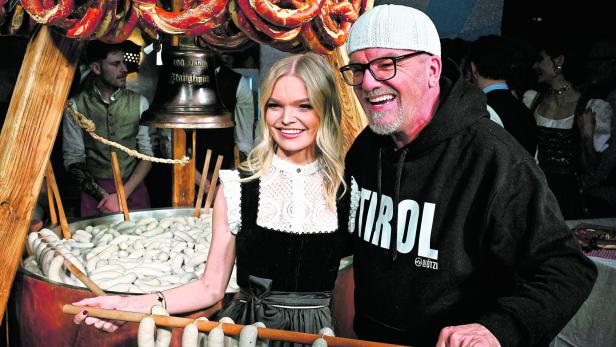 Eine Frau in Dirndl und ein Mann mit Tirol-Pullover halten gemeinsam einen Holzstab mit Weißwürsten, im Hintergrund Brezeln.