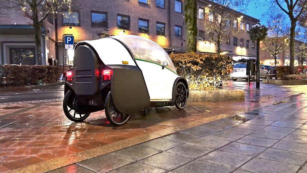 Ein vierrädriges, geschlossenes E-Bike steht auf nassem Gehweg in einer beleuchteten Stadtstraße.