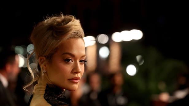 Eine elegant gekleidete Frau mit hochgesteckten blonden Haaren blickt über die Schulter, im Hintergrund unscharfe Lichter.