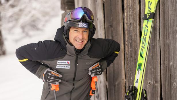 Ein Skifahrer in Winterkleidung lehnt mit Skistöcken an einer Holzwand, neben ihm stehen gelbe Skier.
