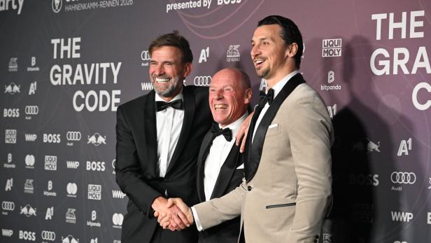 Drei Männer in festlicher Kleidung lachen und posieren gemeinsam vor einer Sponsorenwand.