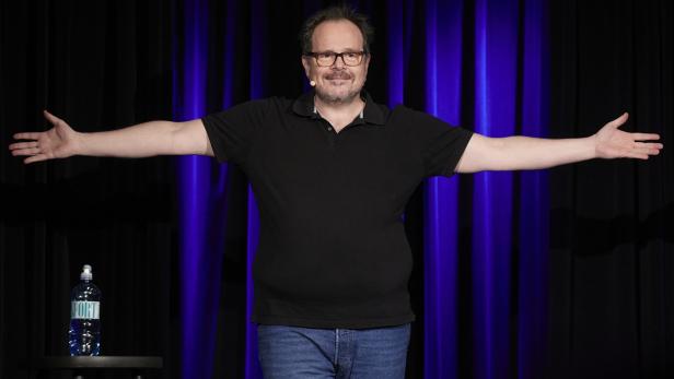 Ein Mann mit Brille und Mikrofon steht mit ausgebreiteten Armen auf einer Bühne vor dunklem Vorhang, daneben ein Hocker mit Wasserflasche.