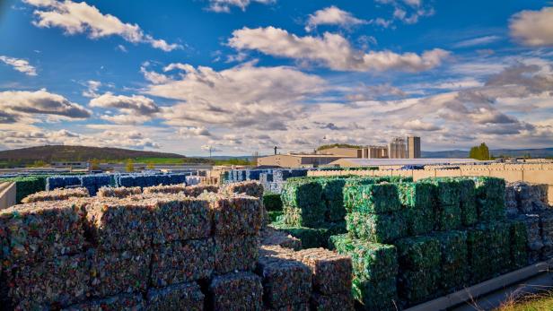 Gestapelte, gepresste Ballen aus recyceltem Kunststoff lagern im Freien vor einer Industrieanlage.