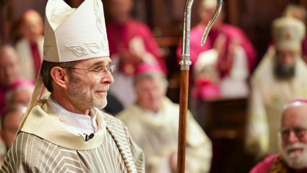 Ein Bischof mit Mitra und Bischofsstab steht vor Geistlichen im festlichen Ornat.