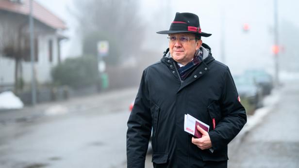 Ein Mann mit Hut und Jacke hält Papiere in der Hand und geht auf einer nebligen Straße.