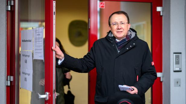 Ein Mann mit Brille und Jacke steht vor einer roten Tür mit Hinweisen zum Wahllokal.