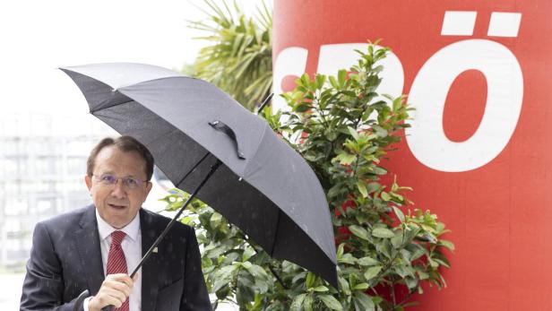 Ein Mann mit Anzug und Krawatte hält einen Regenschirm im Regen vor einem roten Hintergrund mit einem weißen "Ö".