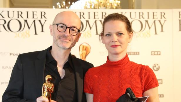 Ein Mann im schwarzen Anzug hält eine goldene Trophäe, neben ihm steht eine Frau in rotem Oberteil und schwarzem Rock.