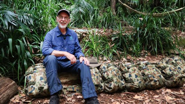 Dr. Bob sitzt in blauer Outdoor-Kleidung im Dschungel neben mehreren Tarnrucksäcken, bereit für die neue Staffel von "Ich bin ein Star – Holt mich hier raus!"
