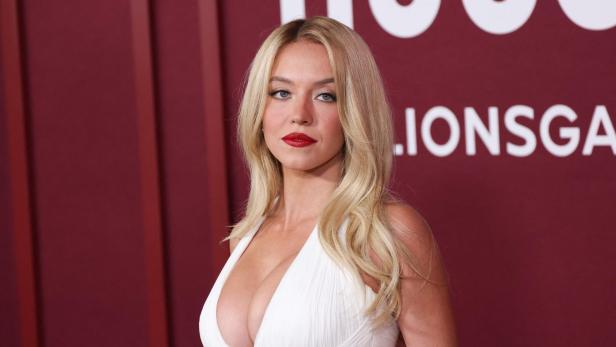 Frau mit langen blonden Haaren und weißem Kleid steht vor einer roten Wand mit weißer Schrift.