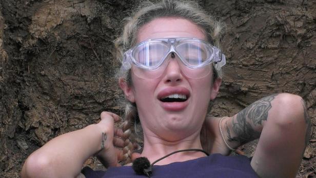 Eine junge Frau mit Schutzbrille und geflochtenen Haaren - Ariel Hediger -wirkt ängstlich und liegt vor einer erdigen Wand.