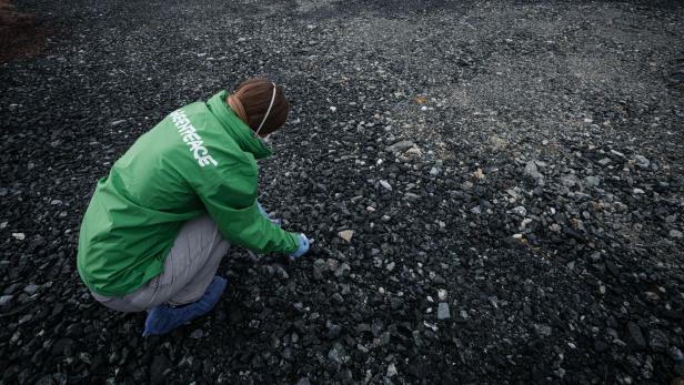 Eine Person mit grüner Jacke und Mundschutz untersucht kniend einen steinigen, grauen Boden auf einer weiten Fläche im Freien.