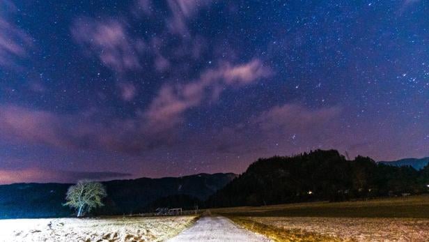 Eine Landstraße führt durch eine nächtliche Landschaft mit Sternenhimmel, Wolken und einem einzelnen Baum am Rand.