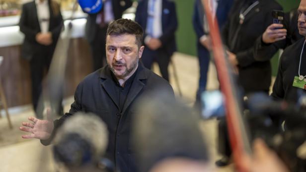 Wolodymyr Selenskyj spricht vor Kameras, umgeben von Sicherheitsleuten und anderen Personen.