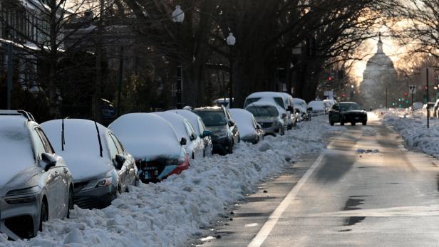 Mehrere Autos sind am Straßenrand geparkt und mit einer dicken Schneeschicht bedeckt, während die Straße geräumt ist.