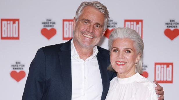 Birgit Schrowange und Ehemann Frank Spothelfer stehen lächelnd Arm in Arm vor einer Wand mit roten Herzen und Logos.