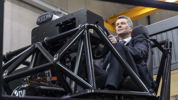 Ein Mann im Anzug sitzt in einem Fahrsimulator mit schwarzem Stahlrahmen und blickt nach oben.