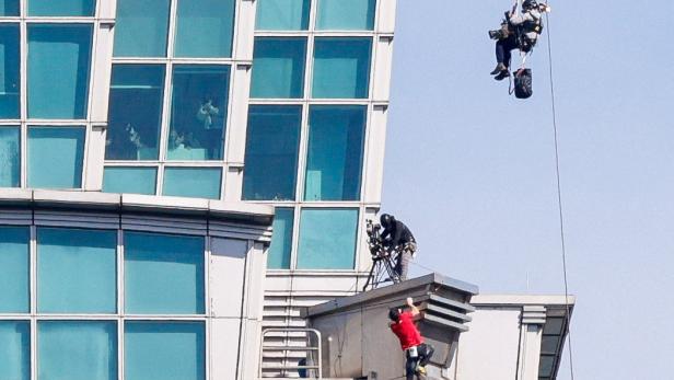 Eine Person klettert an einer großen Metallskulptur an einem Hochhaus, während zwei weitere Personen an Seilen hängen.