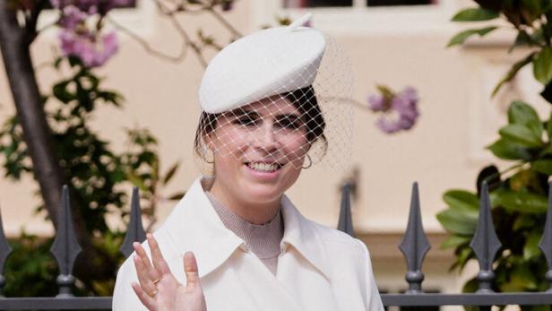 Prinzessin Eugenie mit weißem Mantel und Hut mit Netzschleier lächelt und winkt, in der anderen Hand hält sie eine beigefarbene Tasche.